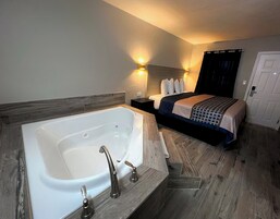 A modern bedroom with a large white bathtub, a bed with a dark headboard, and two wall-mounted lights.