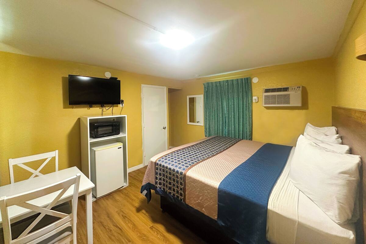 Quarto de hotel com cama, televisão, pequena escrivaninha e guarda-roupa.