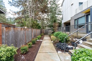 Un étroit sentier mène à un bâtiment, bordé d’une clôture en bois d’un côté et d’une rambarde en métal de l’autre.