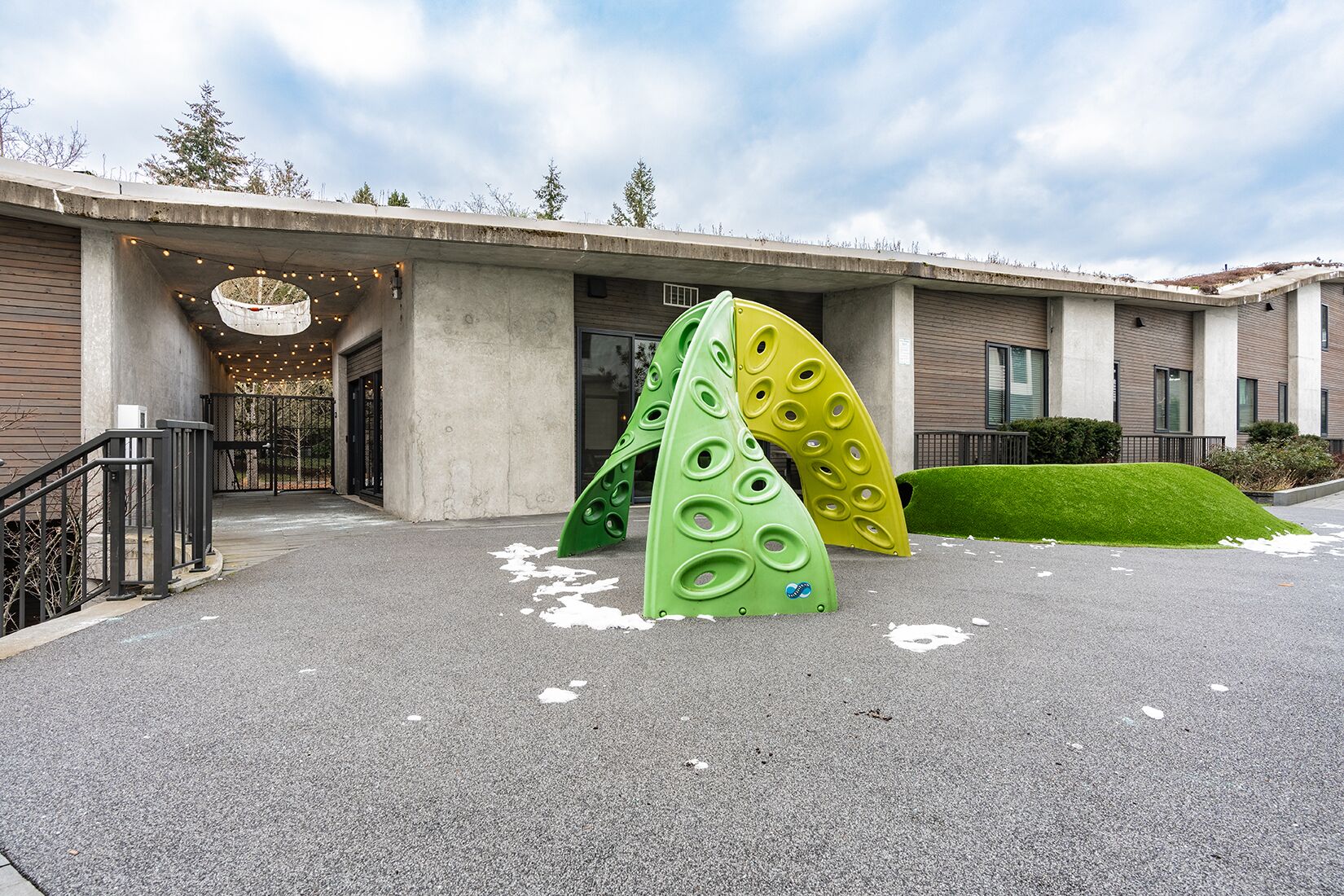A building entrance with a concrete facade, a green and yellow structure resembling a slide, and a small grassy area.