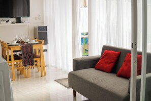 A modern living room with a grey sofa and red cushions, a dining area with wooden chairs and a table, and a large flat-screen TV mounted on the wall. - Otia Villa by Premier Hospitality Asia (Nusa Dua)