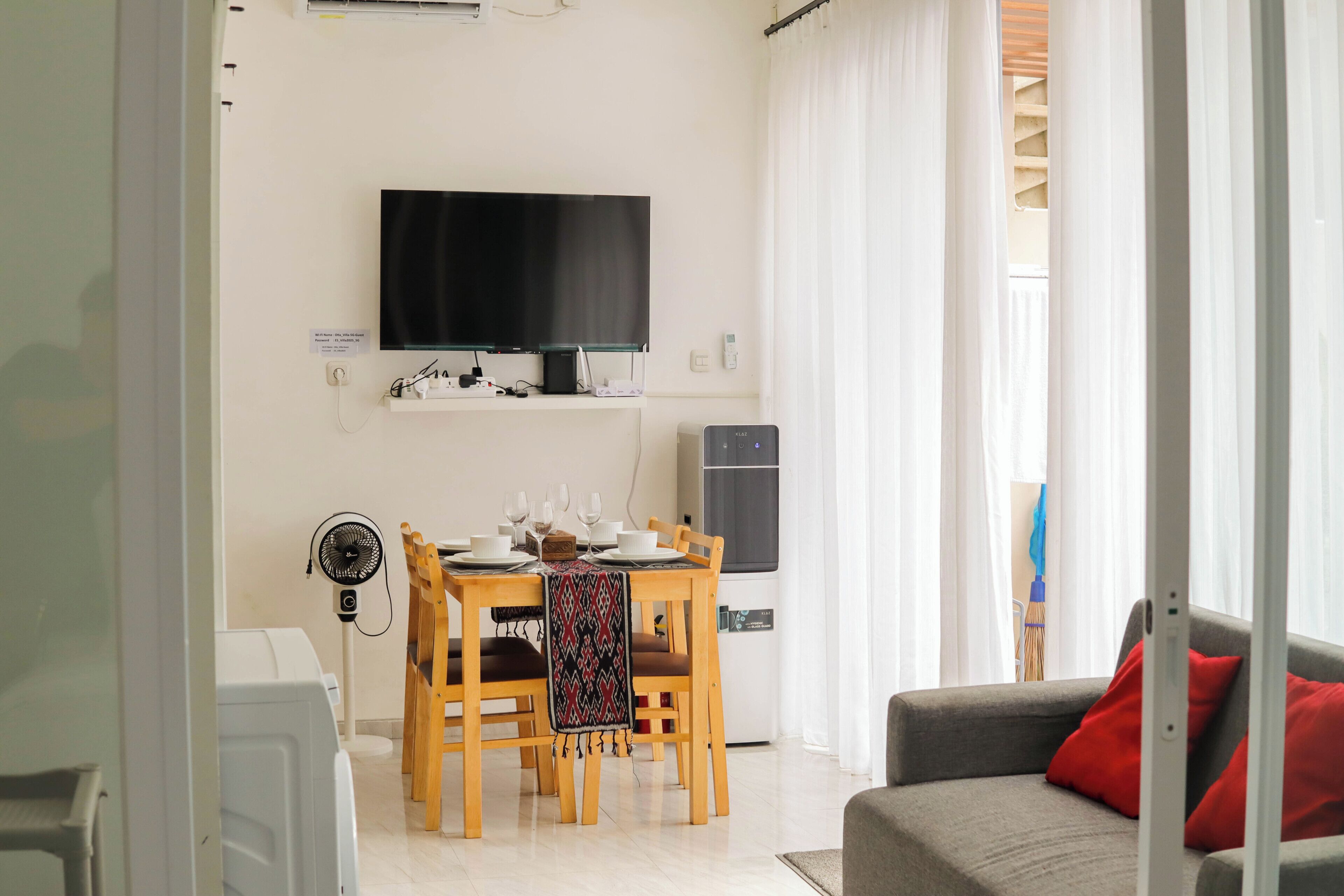 A modern living room with a flat-screen TV mounted on the wall, a dining table set with plates and glasses, a sofa with red cushions, and a standing fan.