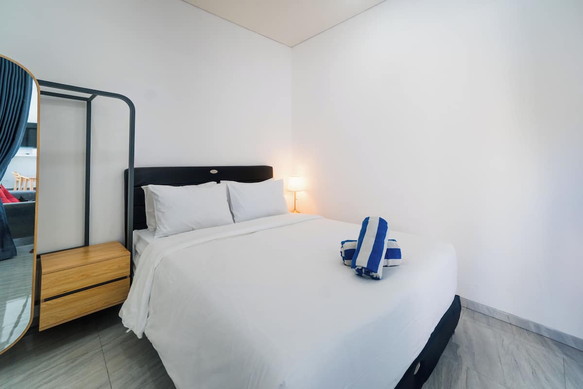 Ein ordentlich gemachtes Bett mit weißen Leinen und einem blau-weiß gestreiften Handtuch, ein Nachttisch mit Lampe sowie ein Holznachtkästchen.