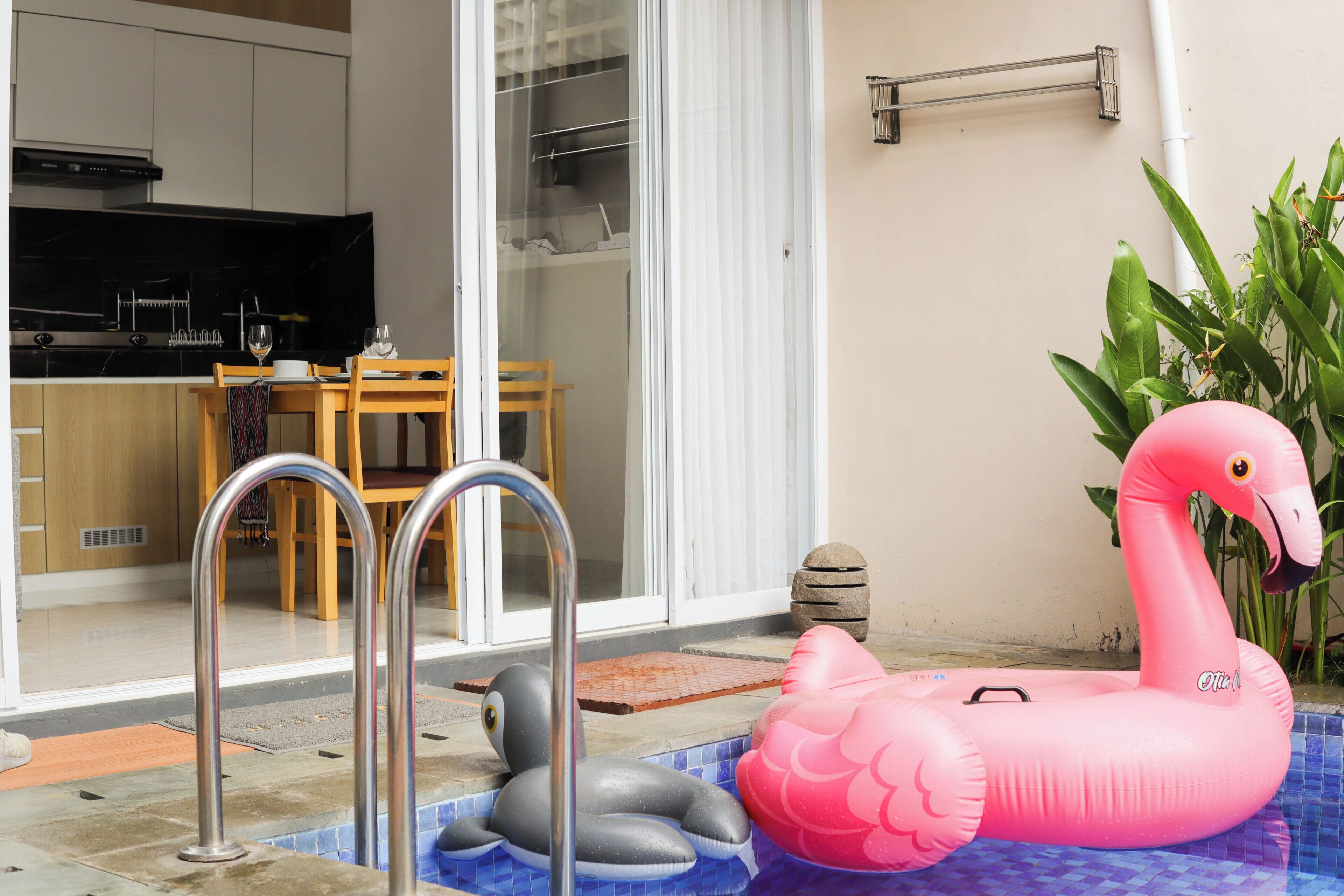 Piscina com boias de flamingo e baleia infláveis, mesa de jantar de madeira com cadeiras e área de cozinha visível através de uma porta de vidro.