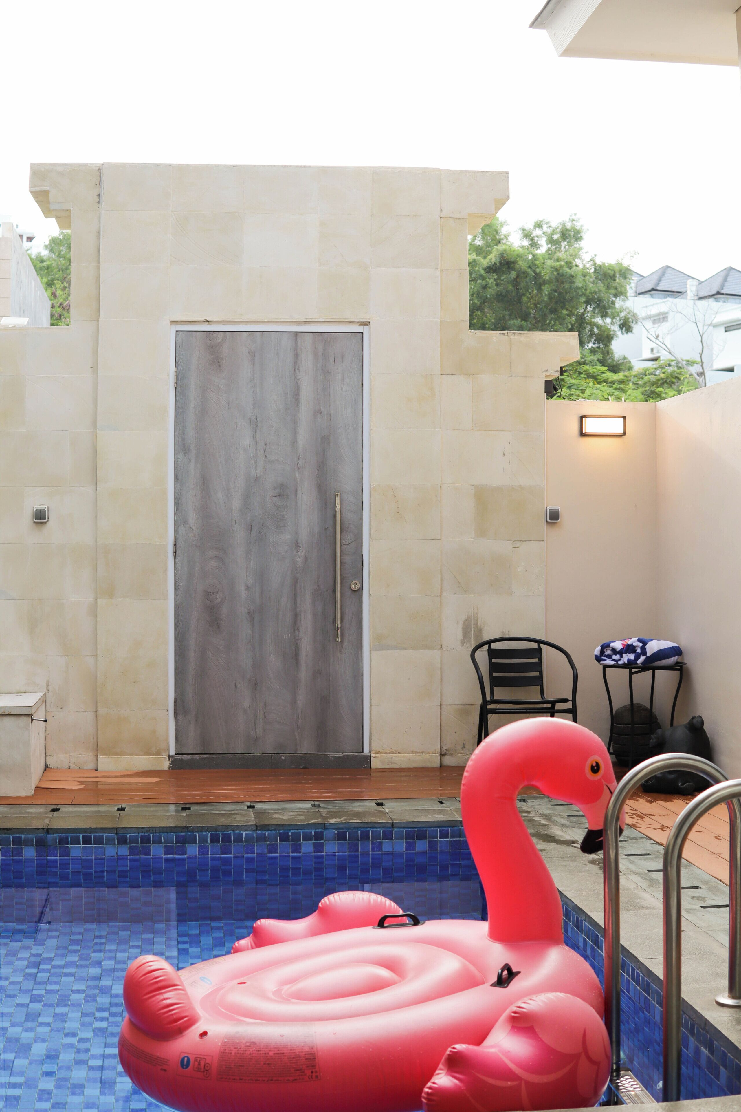 A swimming pool with a pink flamingo float, a door, and a small table with a chair.