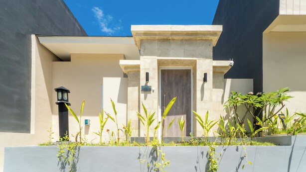 A modern building with a flat roof, a central entrance with a door, and a small garden area with plants and a lamp post. - Otia Villa by Premier Hospitality Asia (Nusa Dua)