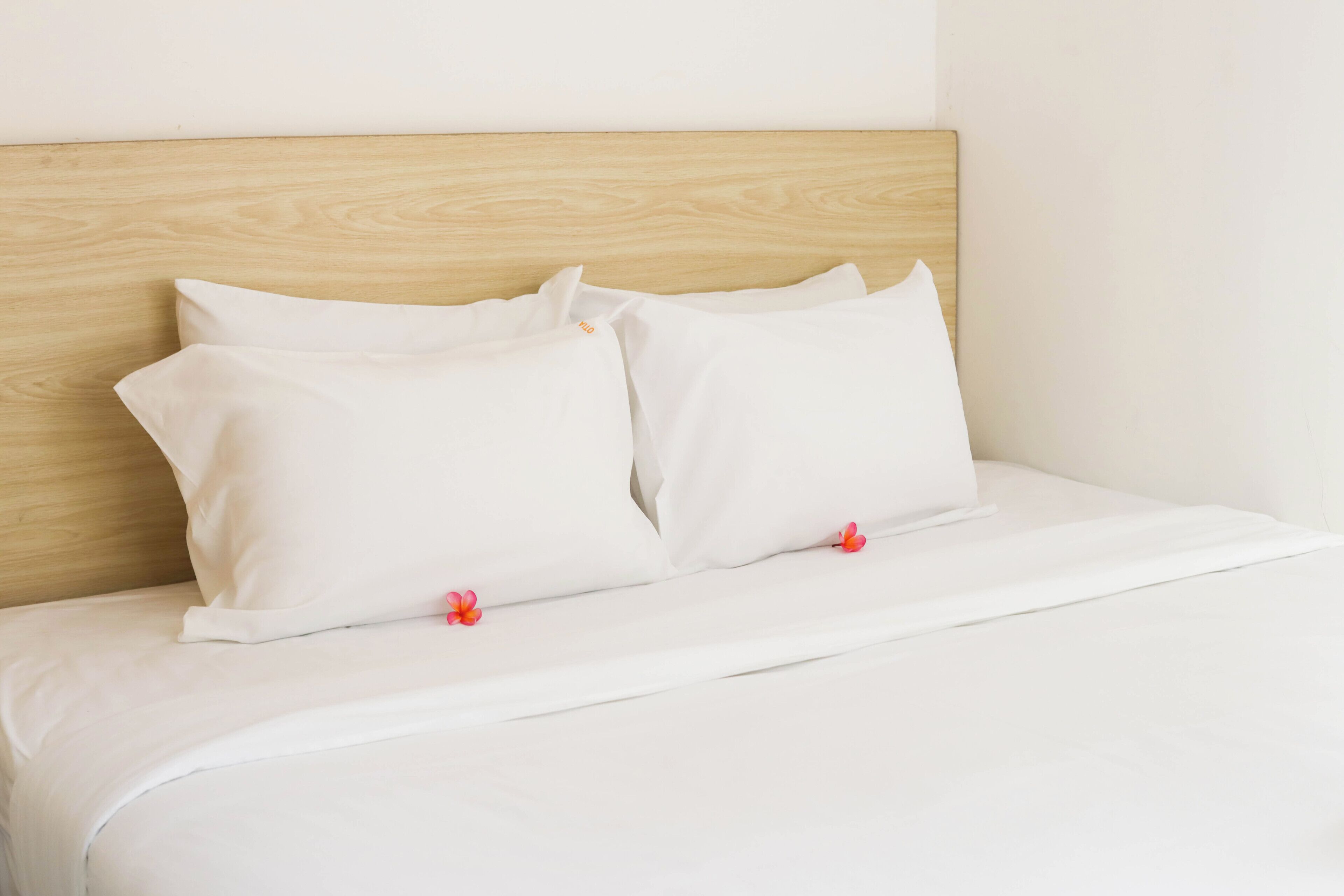 A neatly made bed with white bedding and pillows, a wooden headboard, and a small red flower on the pillow.