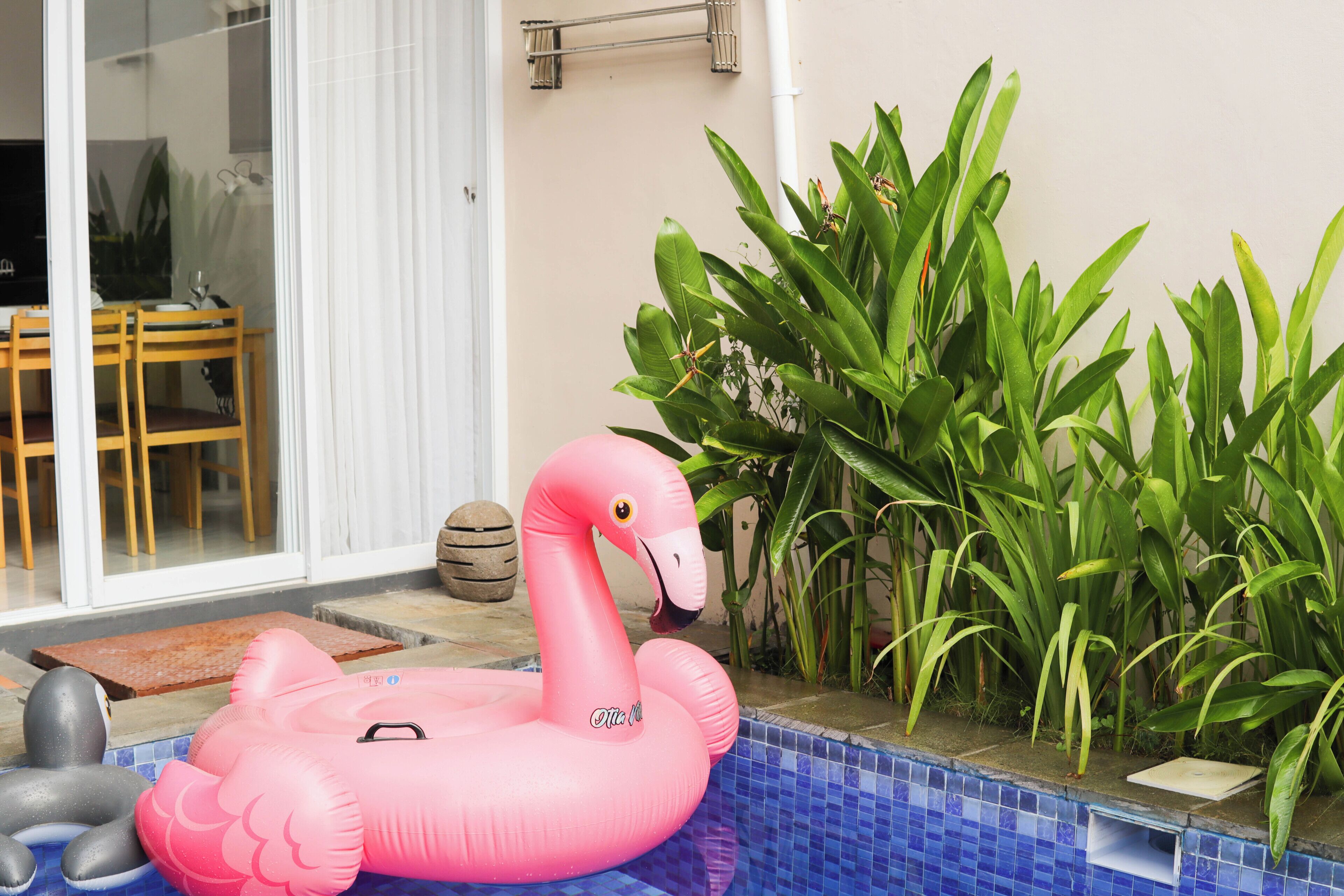 A pink inflatable flamingo by a pool with a tiled edge, surrounded by green plants.