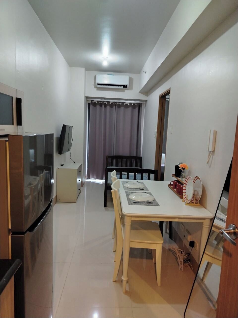 Una cocina compacta con refrigerador, microondas y televisor. Se ve un área de comedor con una mesa y sillas, y una ventana con cortinas al fondo.
