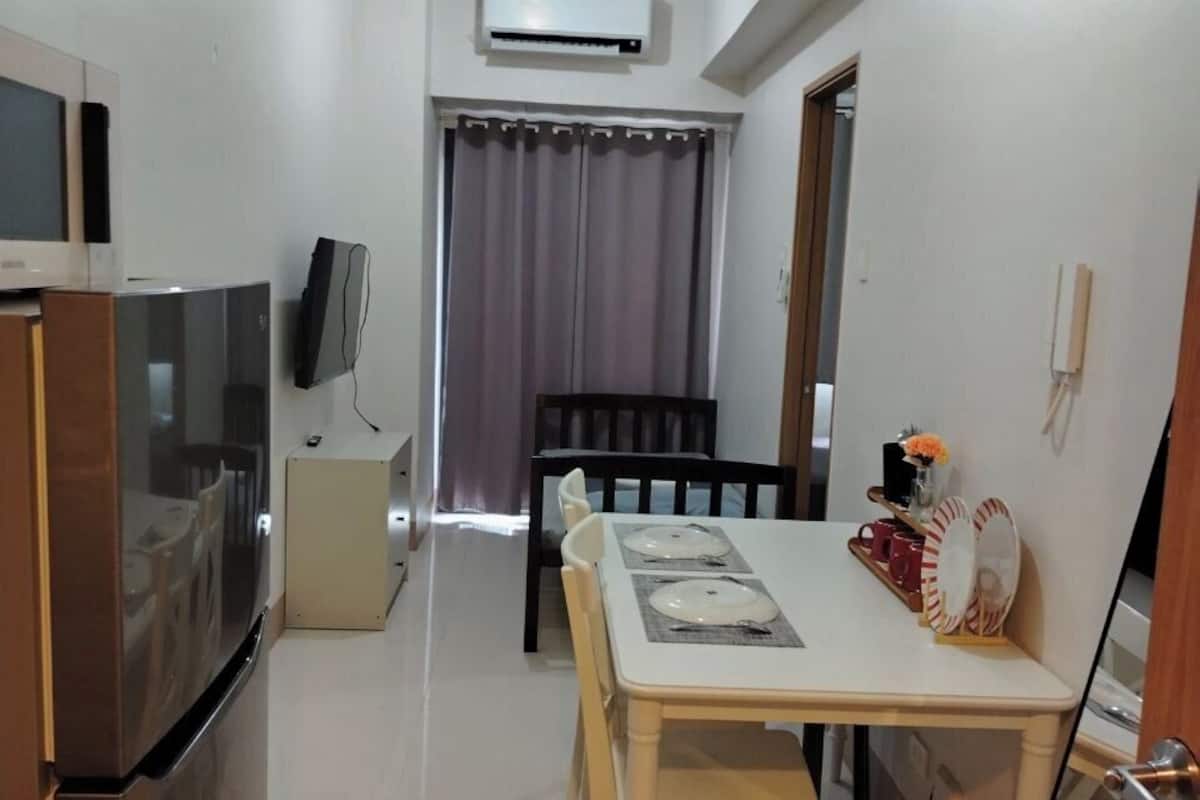 Una cocina compacta con refrigerador, microondas y televisor. Se ve un área de comedor con una mesa y sillas, y una ventana con cortinas al fondo.