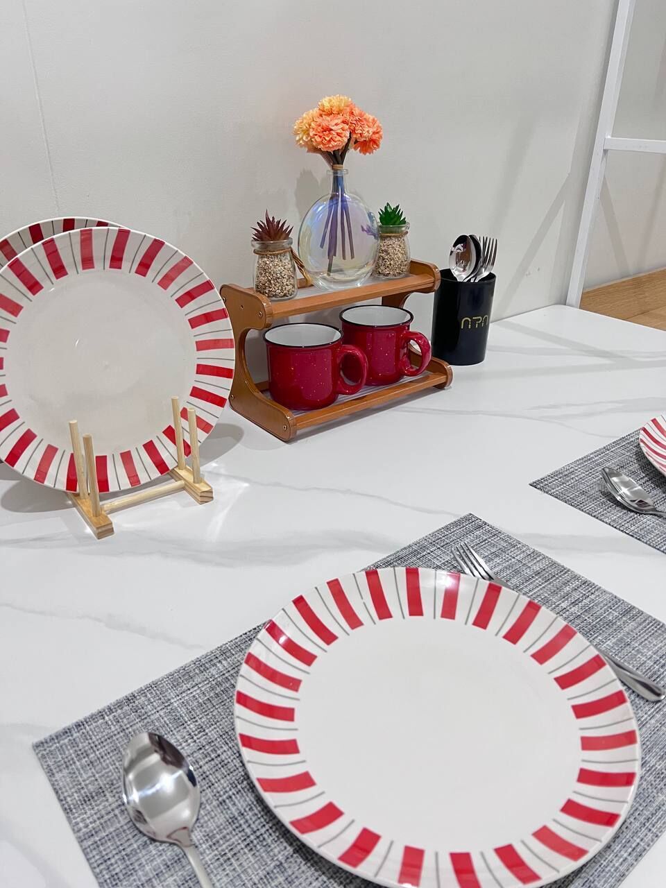 Una mesa de comedor preparada con platos a rayas, tazas rojas y un pequeño jarrón con flores.