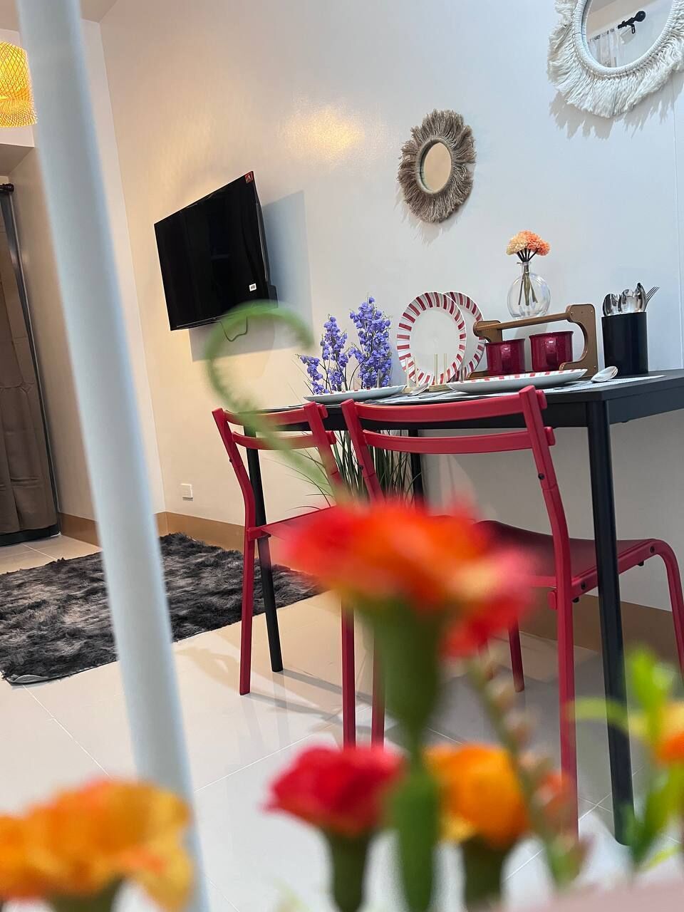 A dining area with a TV mounted on the wall, a table with red chairs, and a decorative mirror.