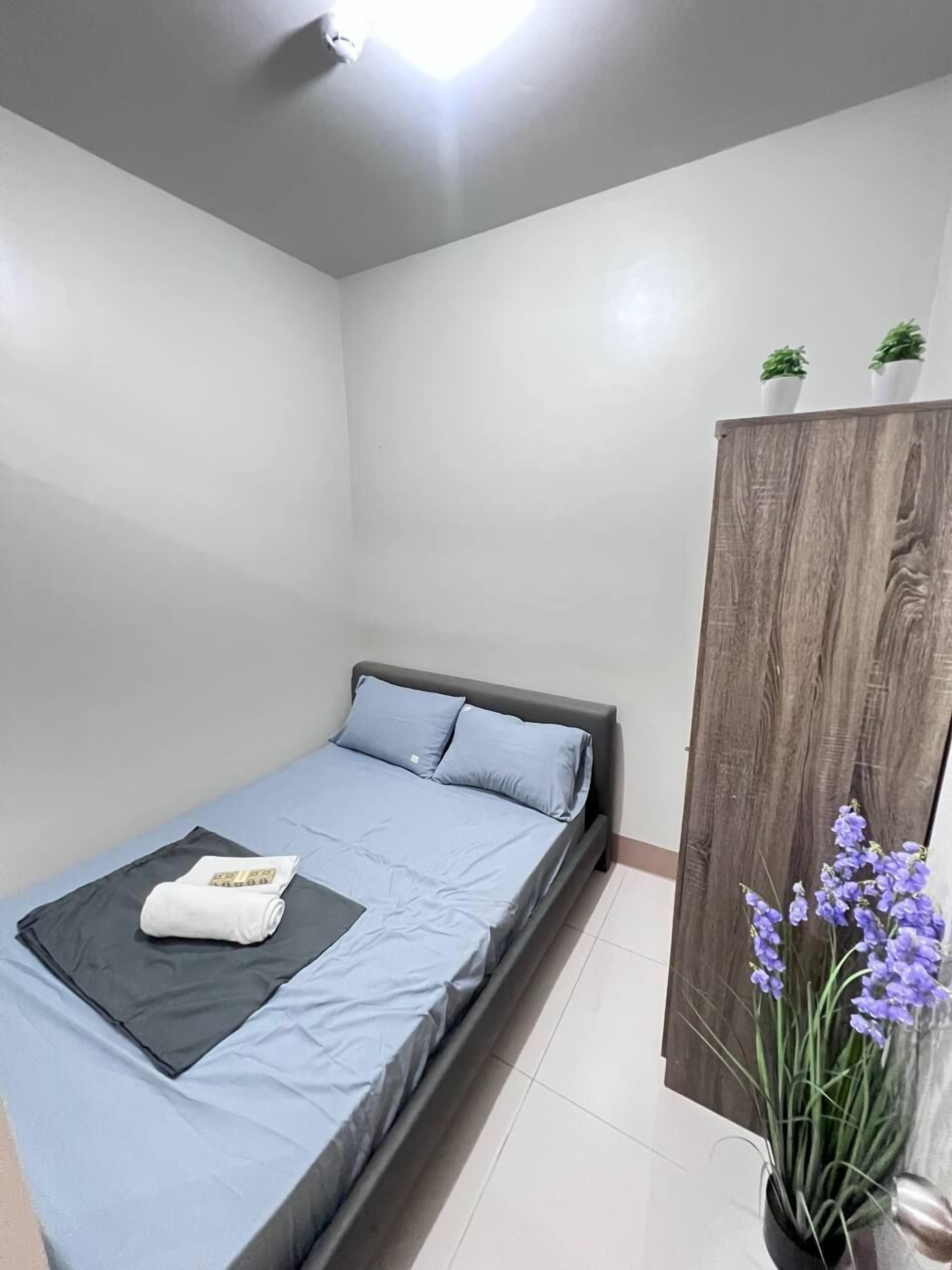 Una cama individual con cabecera gris, colchón azul y manta blanca. Hay una pequeña planta en maceta sobre la cama y otra en un mueble de madera.