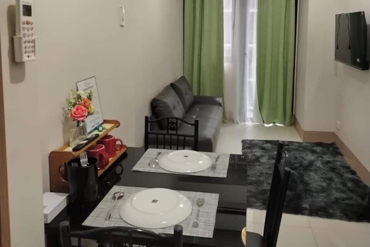 Un coin repas avec une table dressée pour deux personnes, un canapé, un téléviseur fixé au mur et un rideau vert.