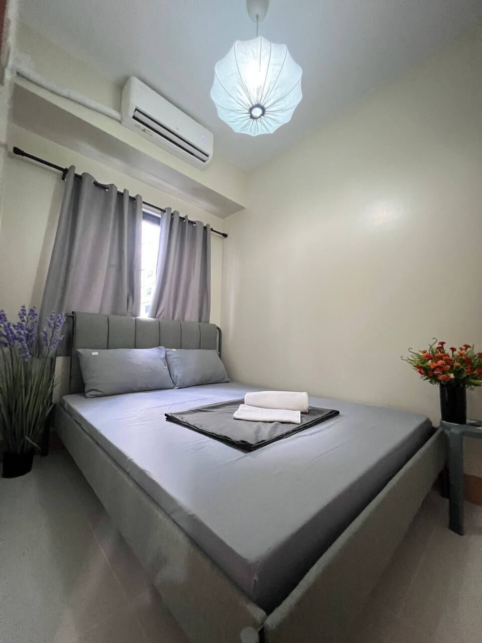 A neatly made bed with a gray mattress and pillows, an air conditioner on the wall, a window with curtains, and a vase with flowers on a nightstand.