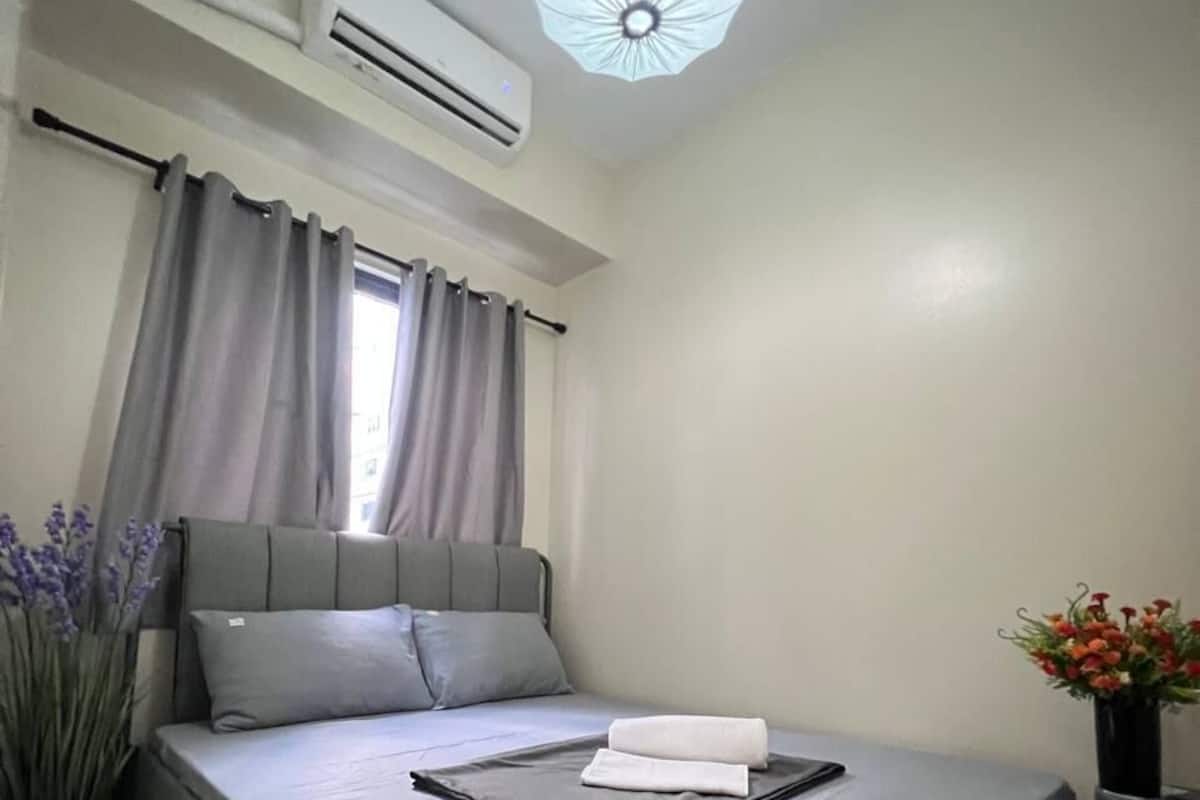 A neatly made bed with a gray mattress and pillows, an air conditioner on the wall, a window with curtains, and a vase with flowers on a nightstand.