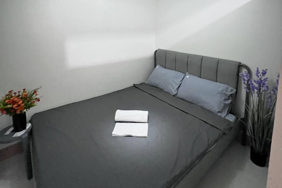 Un letto rifatto con cura, con un materasso e cuscini grigi, un piccolo comodino con un vaso di fiori e una lampada a sospensione.