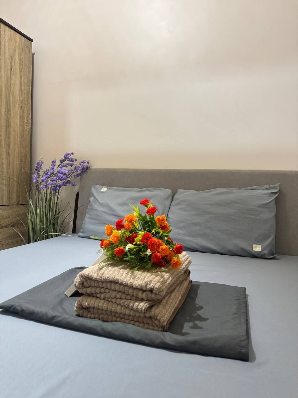 Una cama bien hecha con un colchón y almohadas grises, una pila de toallas color beige y un ramo de flores naranja y rojo.