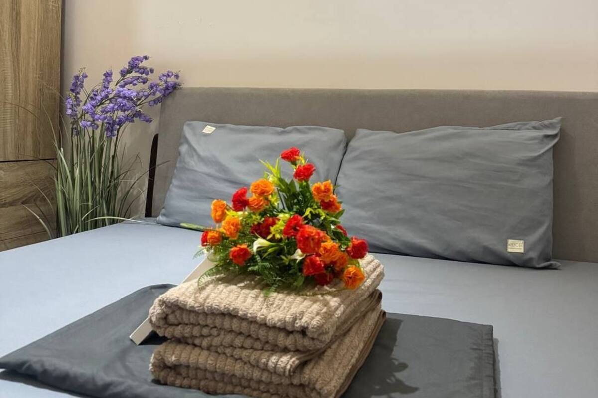 Una cama bien hecha con un colchón y almohadas grises, una pila de toallas color beige y un ramo de flores naranja y rojo.