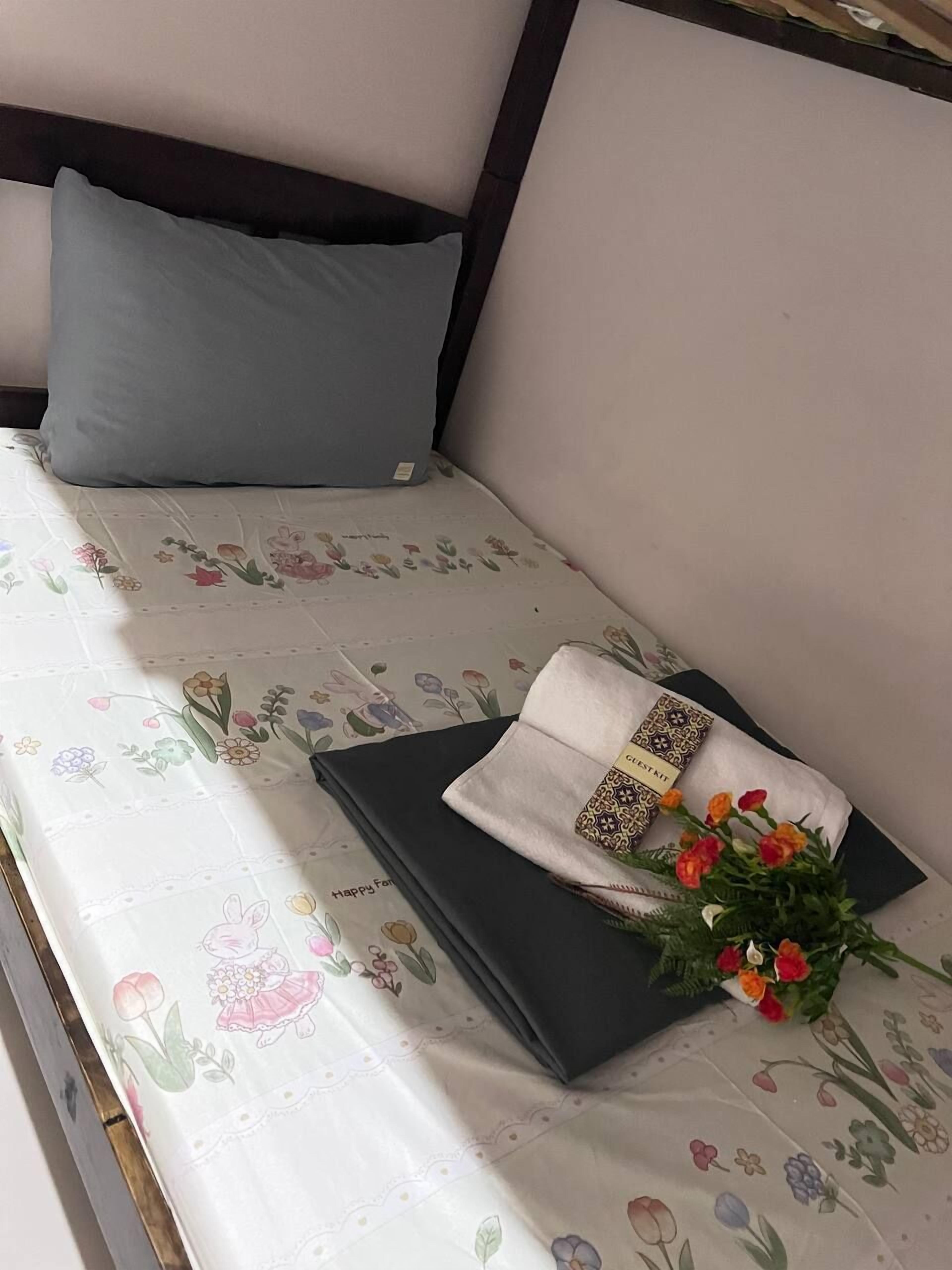 Un letto rifatto con cura, con una coperta a motivi floreali, un cuscino blu e un asciugamano piegato con una passamaneria decorativa.