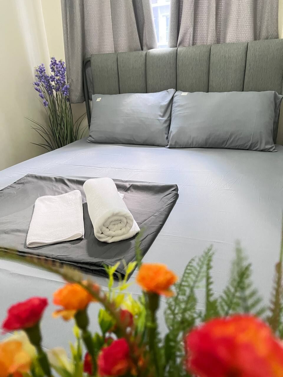 Una cama bien hecha con ropa de cama gris, una toalla blanca enrollada y una toalla blanca doblada sobre la cama.