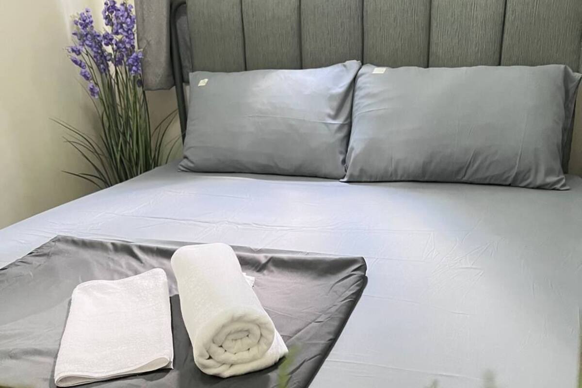 Una cama bien hecha con ropa de cama gris, una toalla blanca enrollada y una toalla blanca doblada sobre la cama.