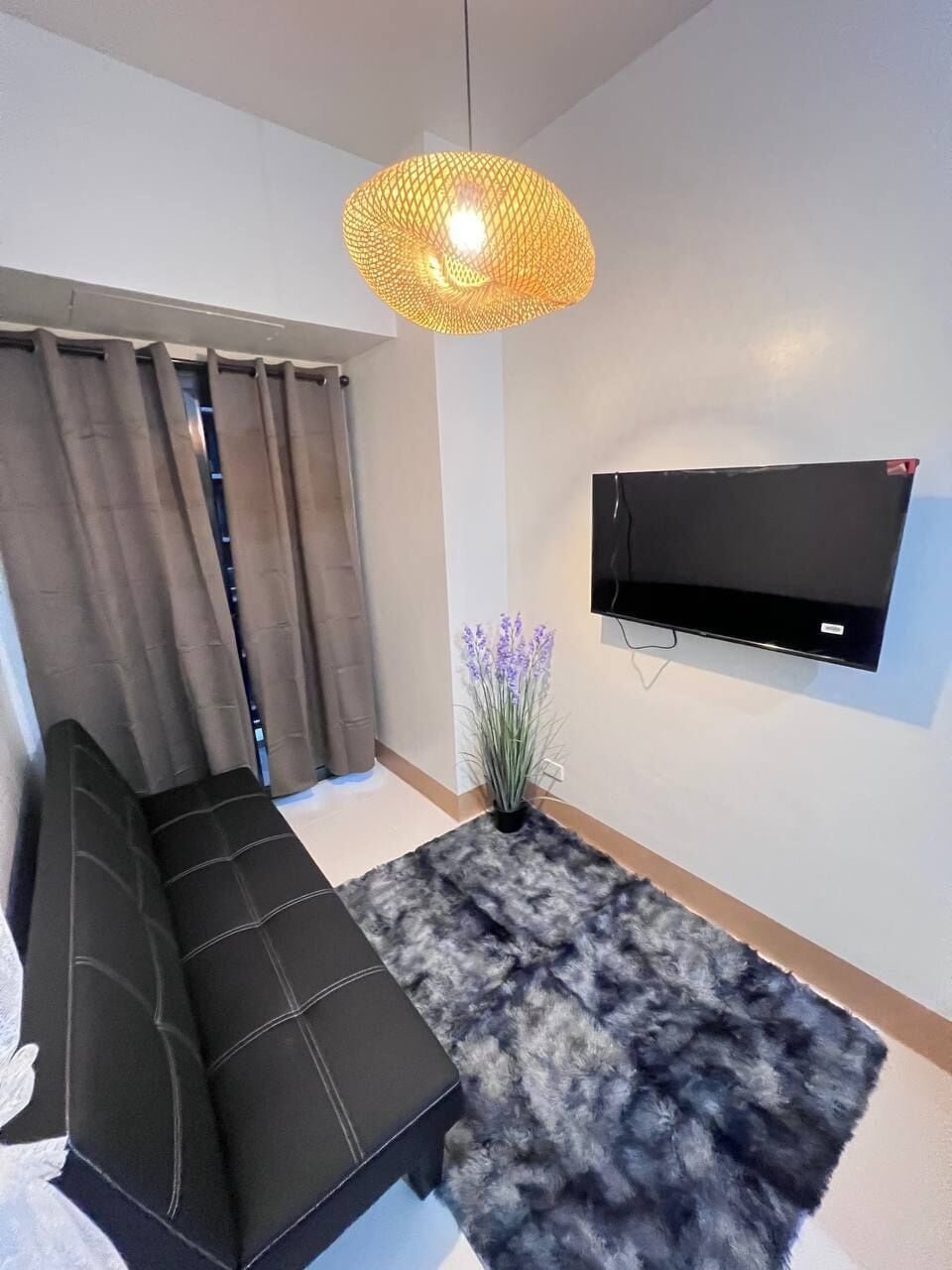 A modern living room with a black sofa, a fluffy rug, a hanging light fixture, a wall-mounted TV, and a window with curtains.