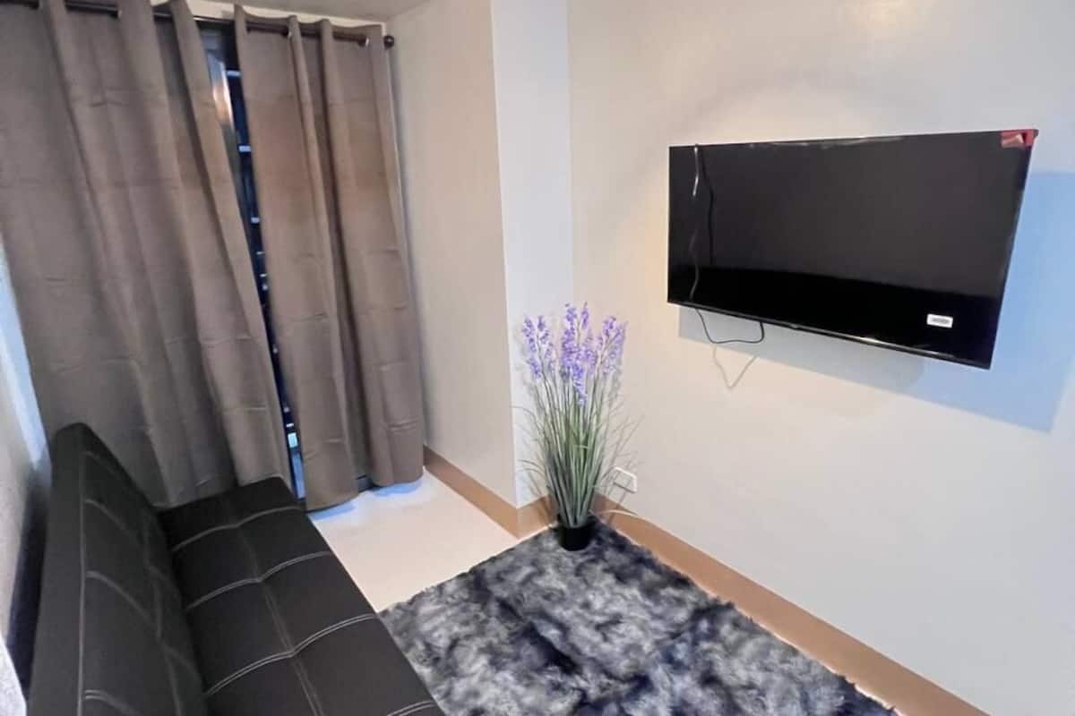 A modern living room with a black sofa, a fluffy rug, a hanging light fixture, a wall-mounted TV, and a window with curtains.