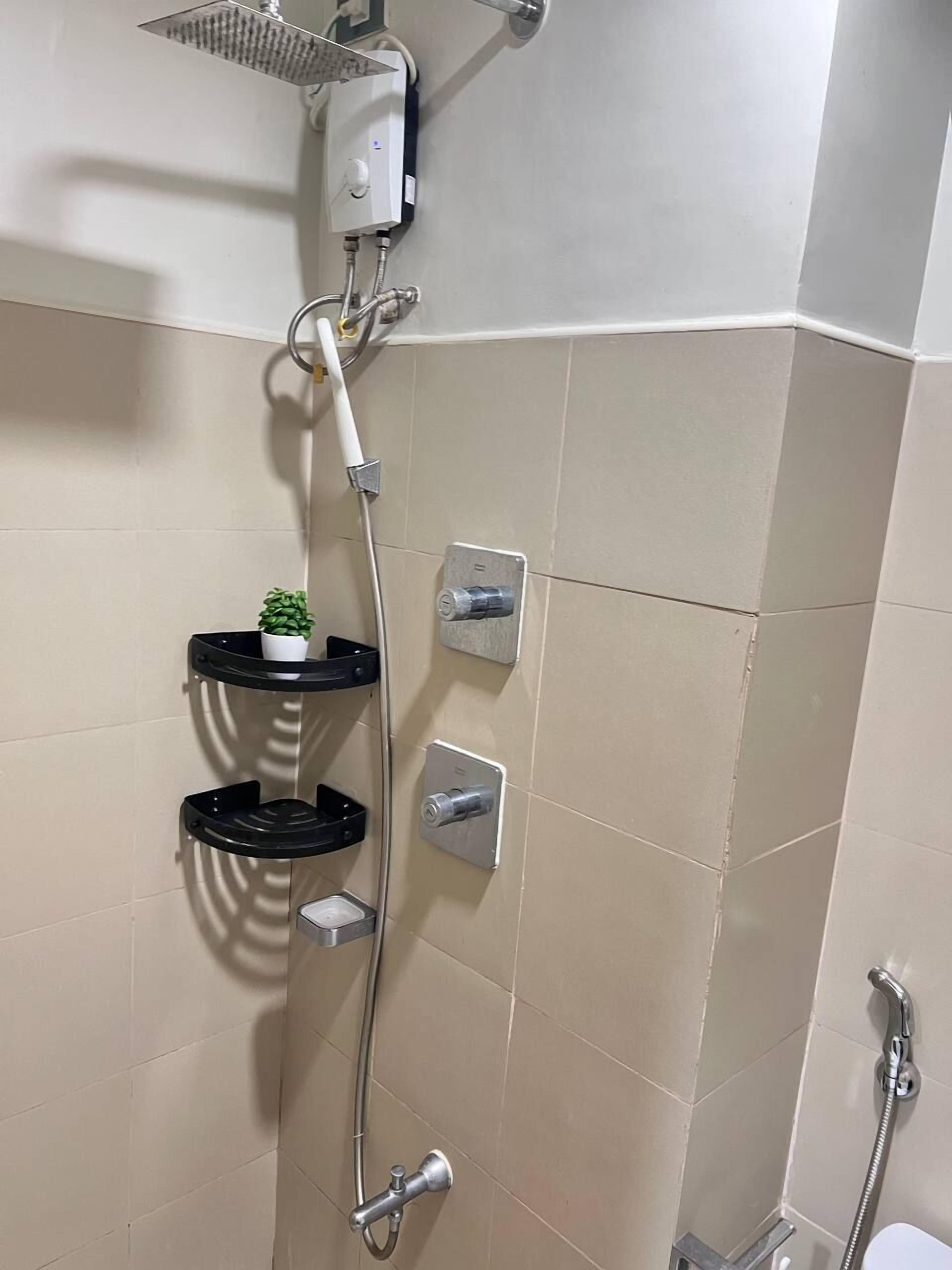 Une douche équipée d’un pommeau de douche à main, d’un panneau de commande fixé au mur et d’une petite étagère sur laquelle est posée une plante.