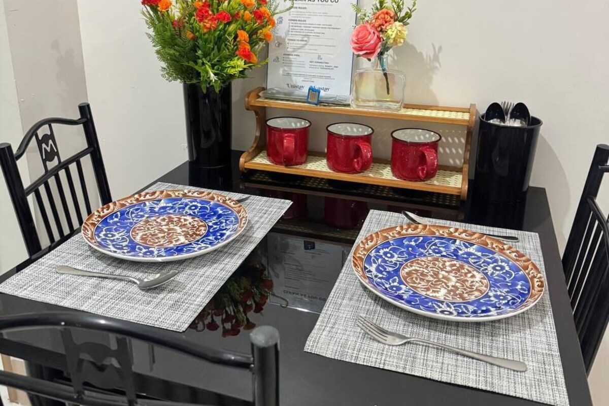 A dining table with two plates, a menu, a vase with flowers, and a set of red mugs.