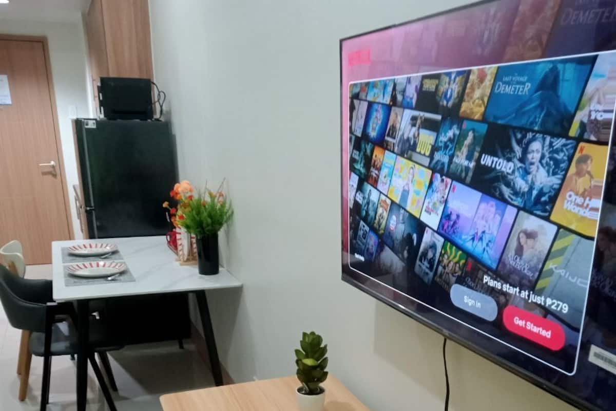Ein modernes Wohnzimmer mit einem an der Wand befestigten Flachbildfernseher, der eine Auswahl an Filmen zeigt. Es gibt einen Esstisch mit Stühlen, einen kleinen Topfpflanzen und einen schwarzen Teppich.