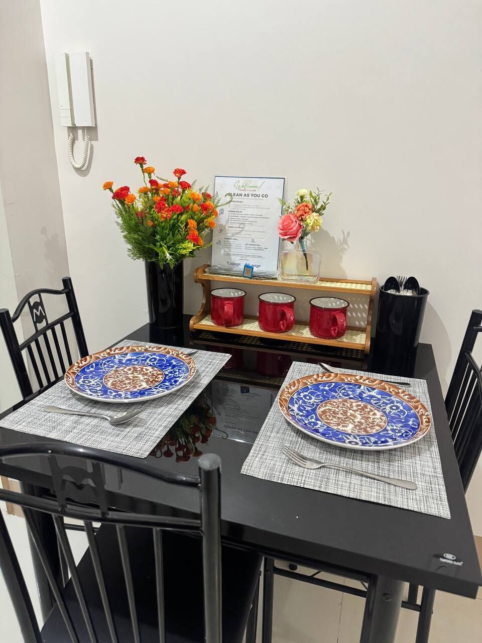 A dining table with two plates, a menu, a vase with flowers, and a set of red mugs.