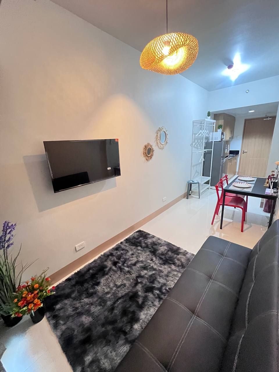 Un salon moderne avec un canapé noir, un téléviseur fixé au mur, un tapis noir et blanc et un coin repas avec des chaises rouges.