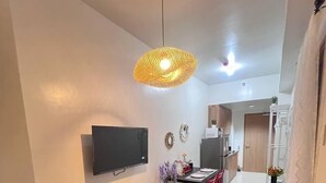 Uma sala de estar moderna com um sofá preto, um tapete estampado, uma TV de tela plana, uma área de jantar com cadeiras vermelhas e uma cozinha ao fundo.