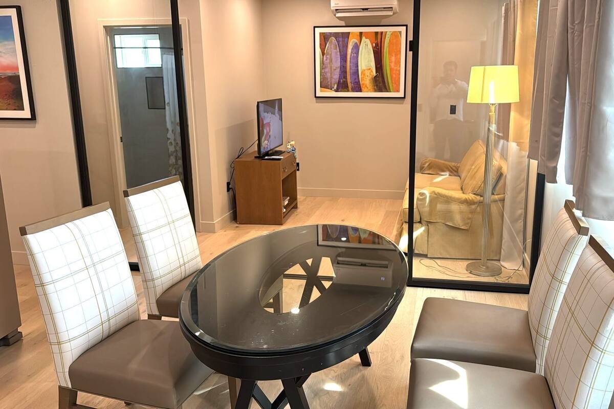 Ein modernes Hotelzimmer mit Glastisch, zwei gepolsterten Sesseln, einer Couch, einem kleinen Holzschrank und einem Bild an der Wand.