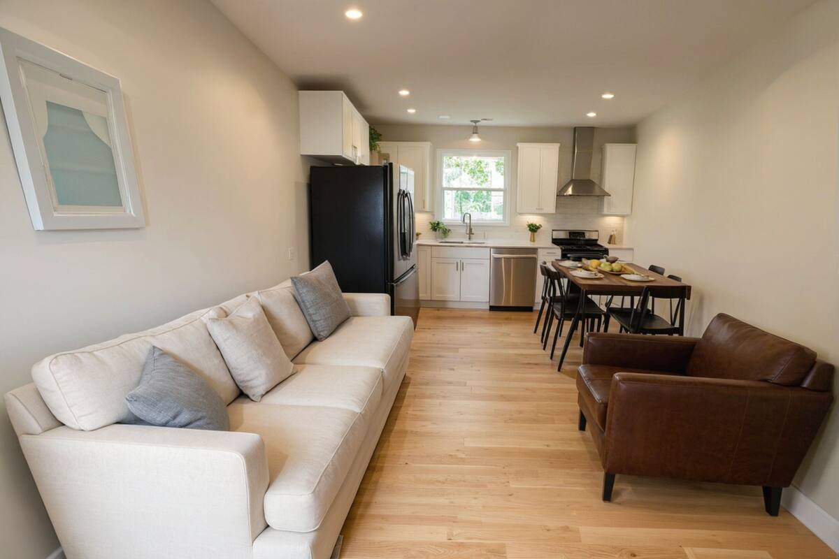 Ein modernes Wohnzimmer mit einer Couch und einem braunen Ledersessel, eine Küche mit weißen Schränken, ein Esstisch und ein Fenster mit Tageslicht.