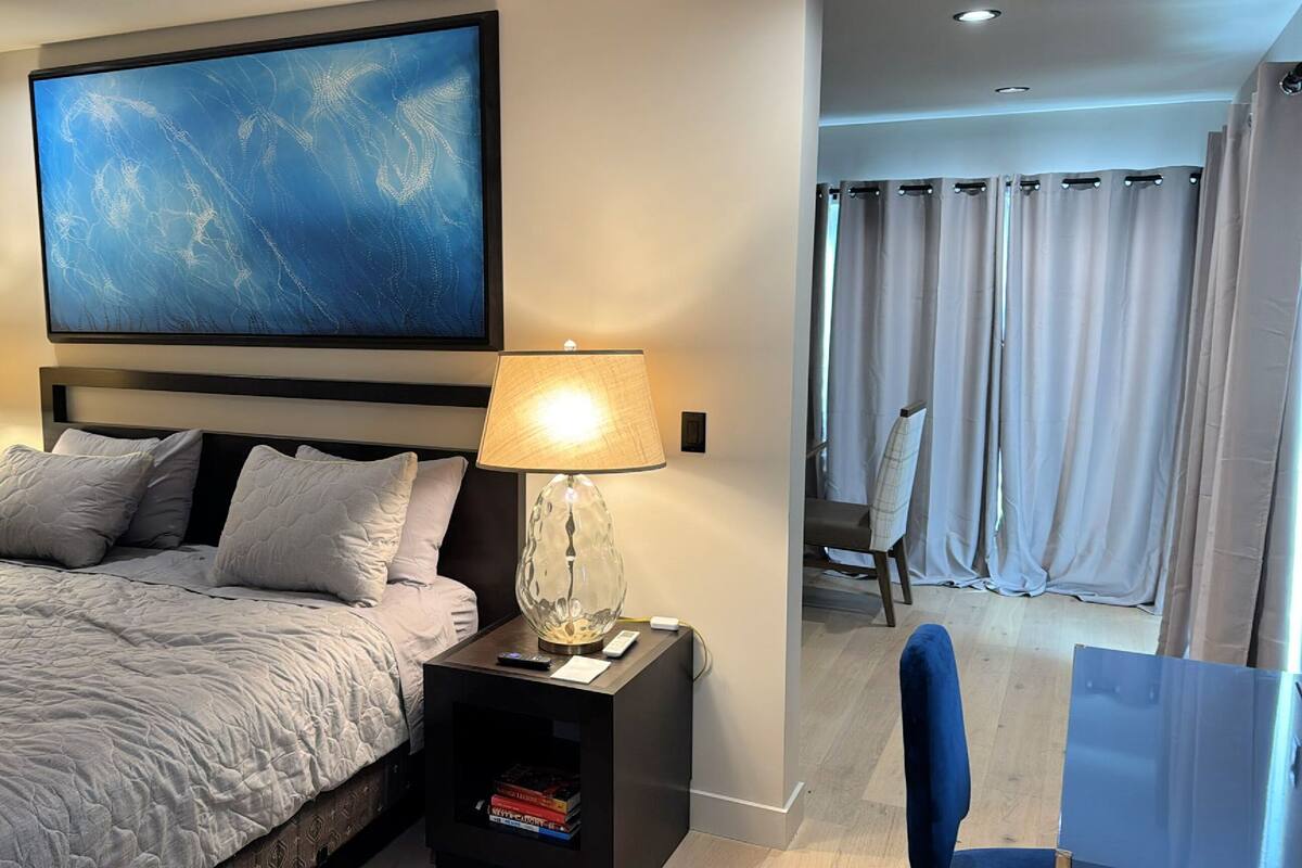 Ein modernes Hotelzimmer mit einem großen Bett, zwei Nachttischlampen, einem Gemälde über dem Bett und einem Blick auf einen Balkon mit Vorhängen.
