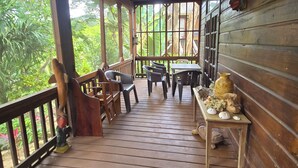 Varanda de madeira com ventilador de teto, cadeiras de plástico, uma mesa com objetos decorativos e vista para vegetação exuberante.