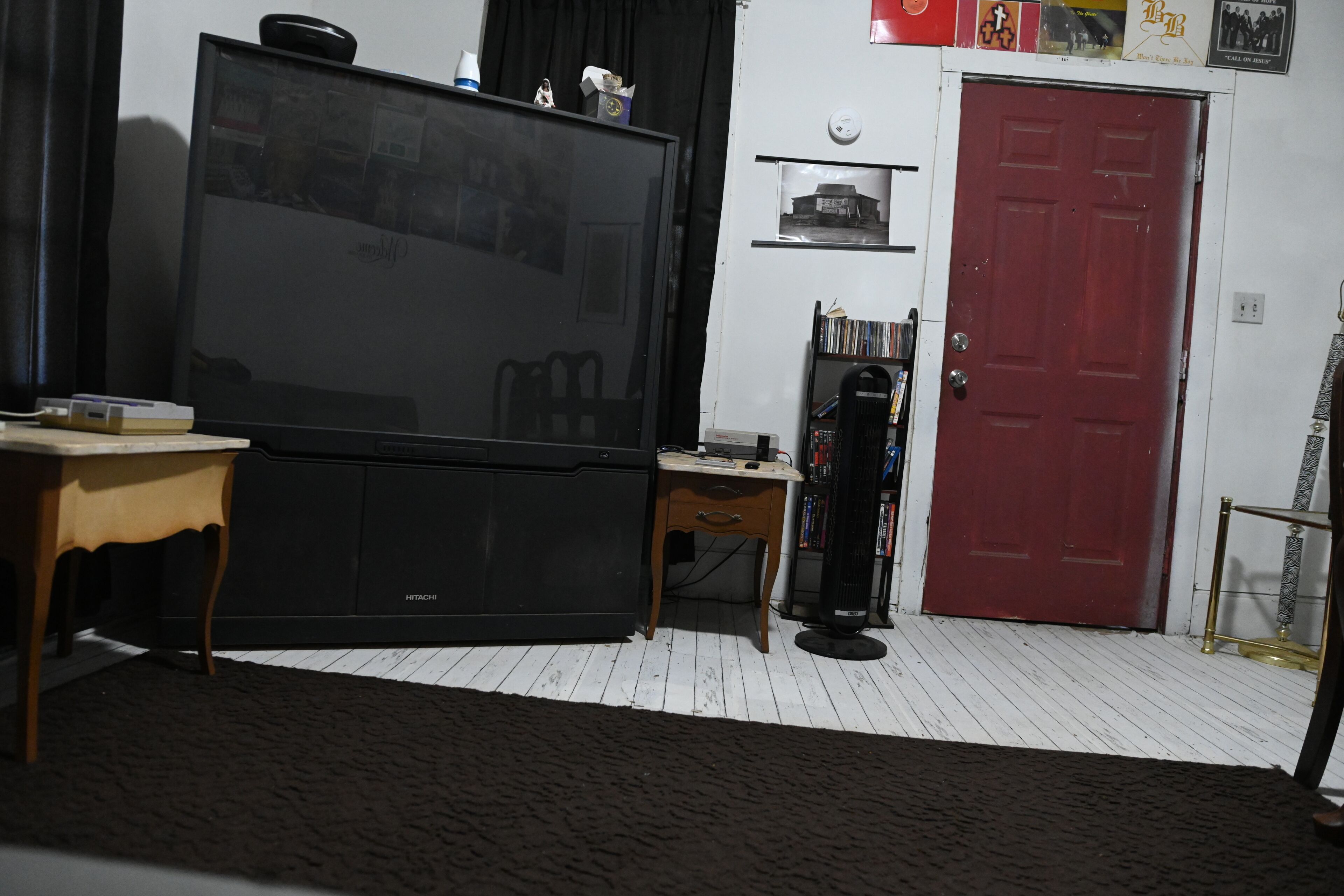 Un salon avec une grande télévision à écran plat, une table en bois, un petit bureau, une bibliothèque, une porte rouge et un tapis à motifs.