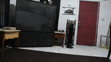 Un salon avec une grande télévision à écran plat, une table en bois, un petit bureau, une bibliothèque, une porte rouge et un tapis à motifs.