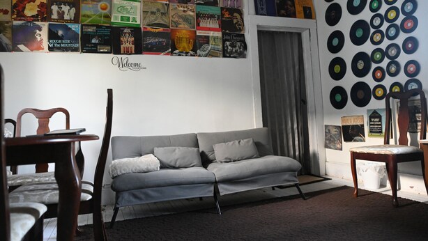 Una habitación con un sofá gris, una silla de madera, una pared decorada con portadas de discos y una pared llena de discos de vinilo.