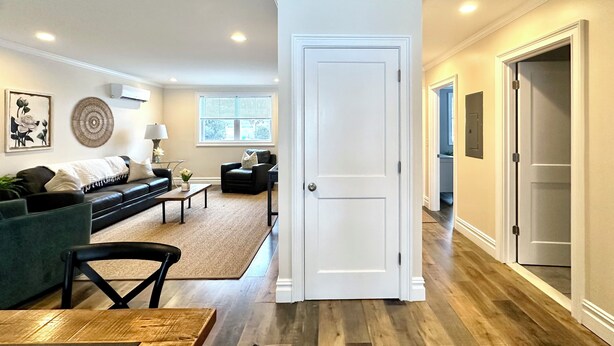 Amplia sala de estar con un sofá verde oscuro, una mesa de centro y una mesa de comedor. Hay un ventanal con persianas, una puerta que conduce a otra habitación y un aire acondicionado instalado en la pared.
