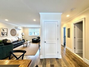 A spacious living room with a dark green sofa, a coffee table, and a dining table. There is a large window with blinds, a door leading to another room, and a wall-mounted air conditioner.