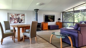 A modern living room with a dining area, a fireplace, a television, and large windows offering a view of the outdoors.