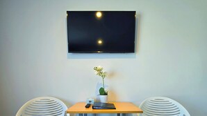 A small room with a TV mounted on the wall, two white chairs, a small table, and a single flower vase. - Kingfisher Lodge Studio (Lake Tekapo)