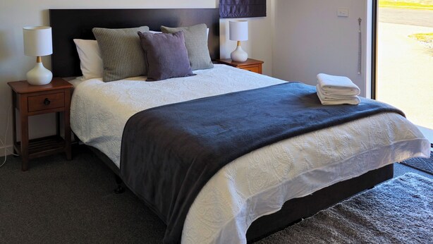 A bedroom with a large bed, two bedside tables, a nightstand, a window with a view, and a painting on the wall. - Kingfisher Lodge Studio (Lake Tekapo)