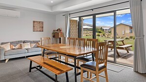 A modern living room with a wooden dining table, grey sofa, and a view of the outdoors through large sliding glass doors.