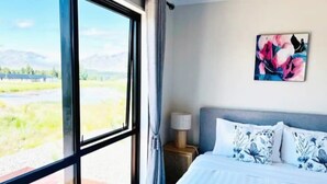 Een slaapkamer met een groot raam, een bed met bloemenkussens, een nachtkastje en een schilderij aan de muur.