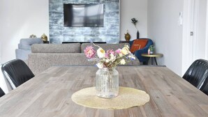 Een eettafel met een bloevas, een geweven placemat, en een modern zithoek met een bank, een televisie en decoratieve objecten.