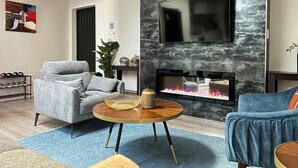 Een moderne woonkamer met een grijze bank, een blauwe fauteuil, een ronde salontafel en een haard met een televisie erboven.
